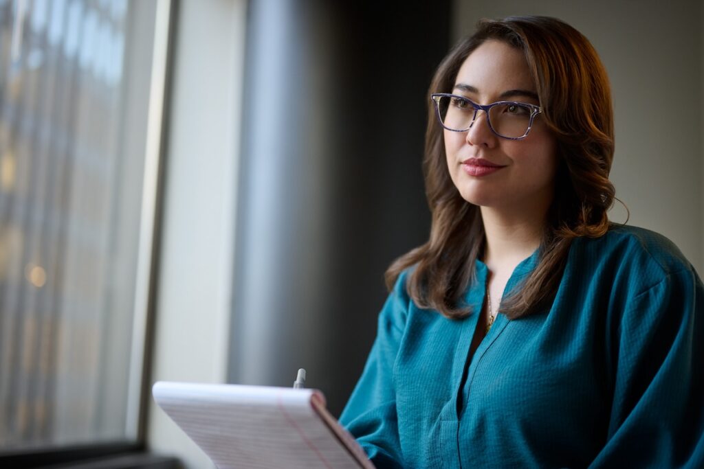 Woman wearing glasses gazing out the window holding a notepad and a pen.